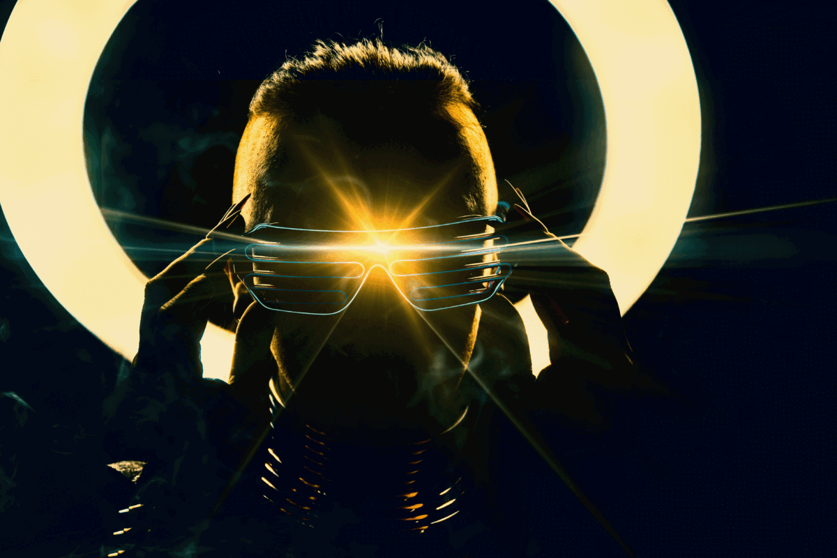 A person in shadow wearing futuristic looking glasses with a bright lens flair emanating from them.