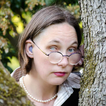 A photo of Elsa Sjunneson, a white woman with dark hair, an occluded cataract on her right eye, and a purple hearing aid, looking at the camera from behind a tree.